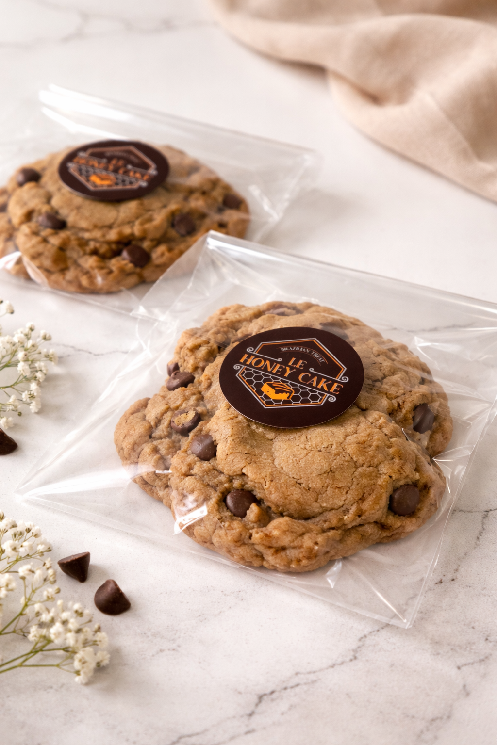 Two cookies with a 'The Honey Cake' label on a marble surface with a beige cloth and flowers in the background.